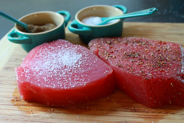 Two raw tuna steaks seasoned with salt and pepper on a wooden cutting board with two small blue salt and pepper containers in the background.