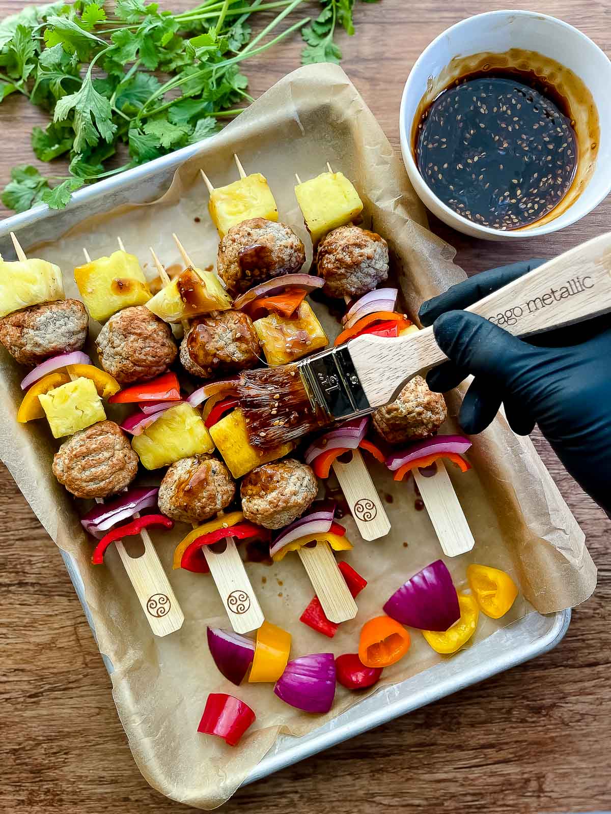 A gloved hand basting turkey meatball skewers with teriyaki sauce placed on top of a baking sheet with a white bowl of sauce on the side and fresh cilantro in the back.