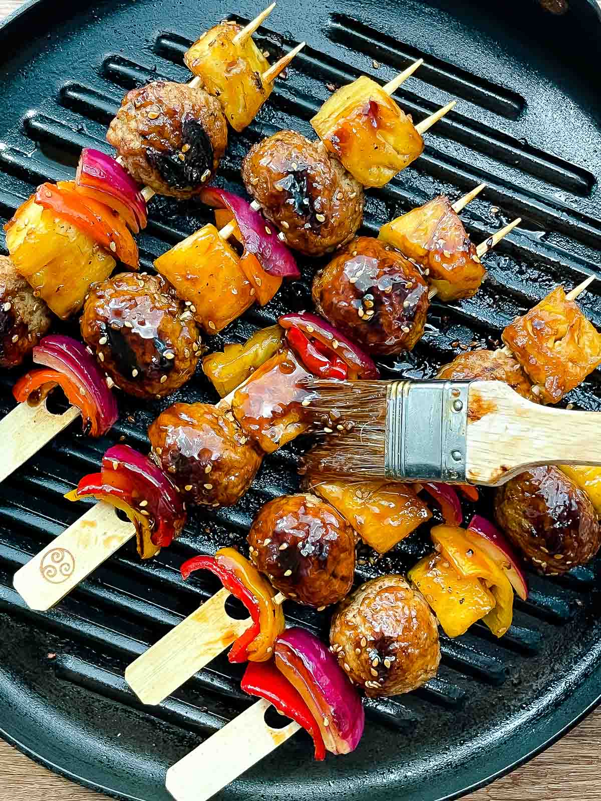 A basting brush basting turkey meatball skewers with teriyaki sauce on top of a stove-top grill pan.