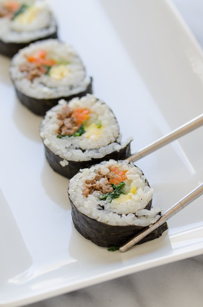 Chopsticks grabbing a piece of ground beef Gimbap or Korean sushi on a white plate.