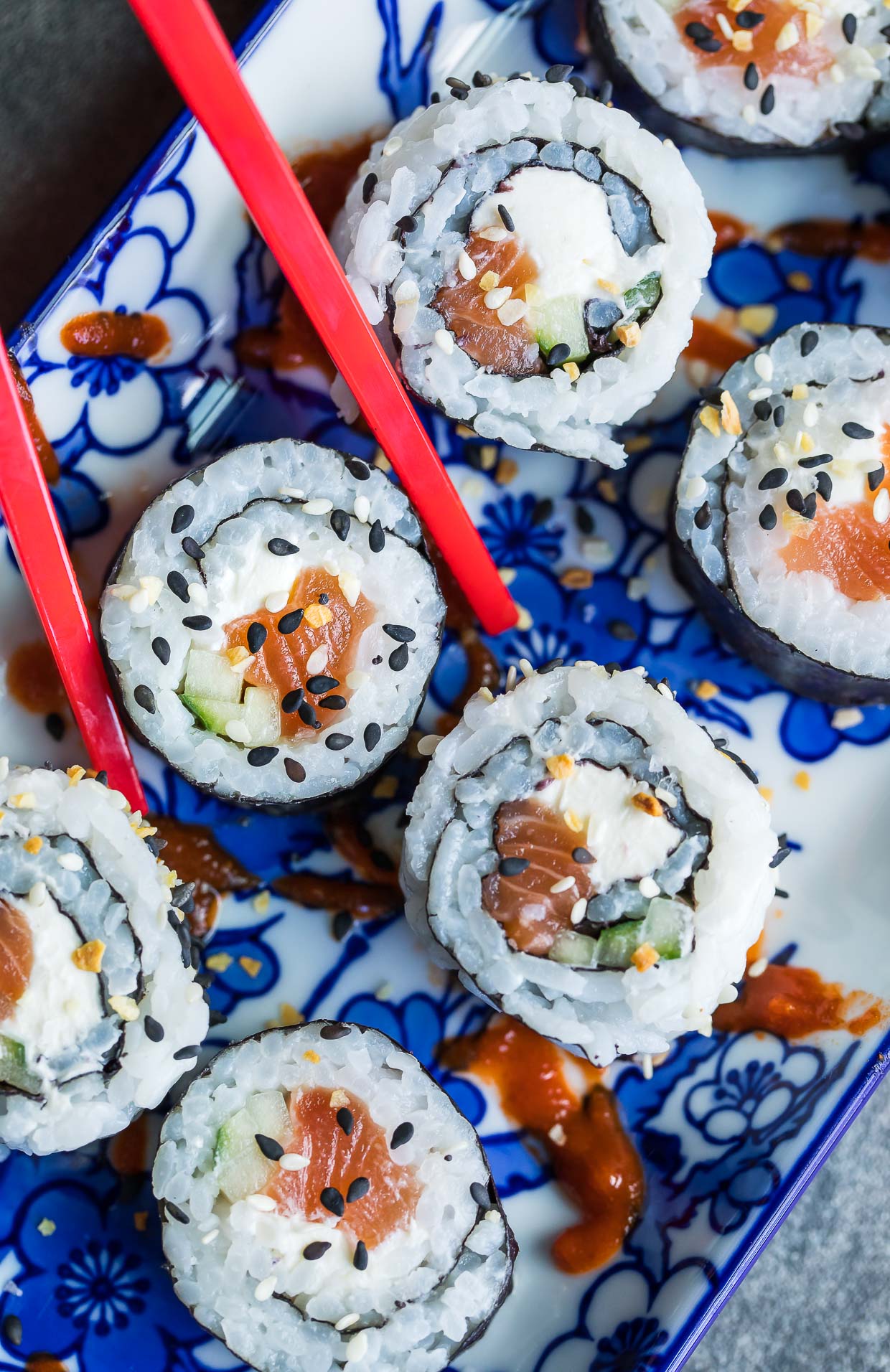 A pair of red chopsticks picking up a piece of Philadelphia sushi roll on a colorful blue and white plate.