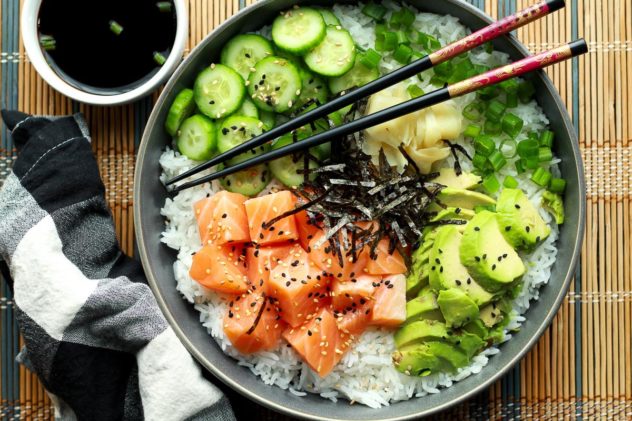 Salmon sushi bowl featuring chunks of sushi-grade salmon, sushi rice, sliced avocados, cucumbers, and seaweed strips on a bamboo mat with chopsticks.