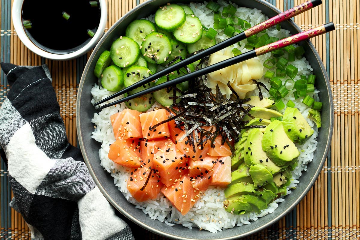 Salmon sushi bowl, healthy salmon recipe, featuring cubed sushi-grade salmon, sushi rice, sliced avocados, cucumbers, and seaweed strips on a bamboo mat with chopsticks.