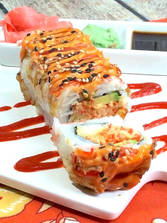 A shaggy dog sushi roll on top of a white cutting board with ginger, wasabi and soy sauce in the background.