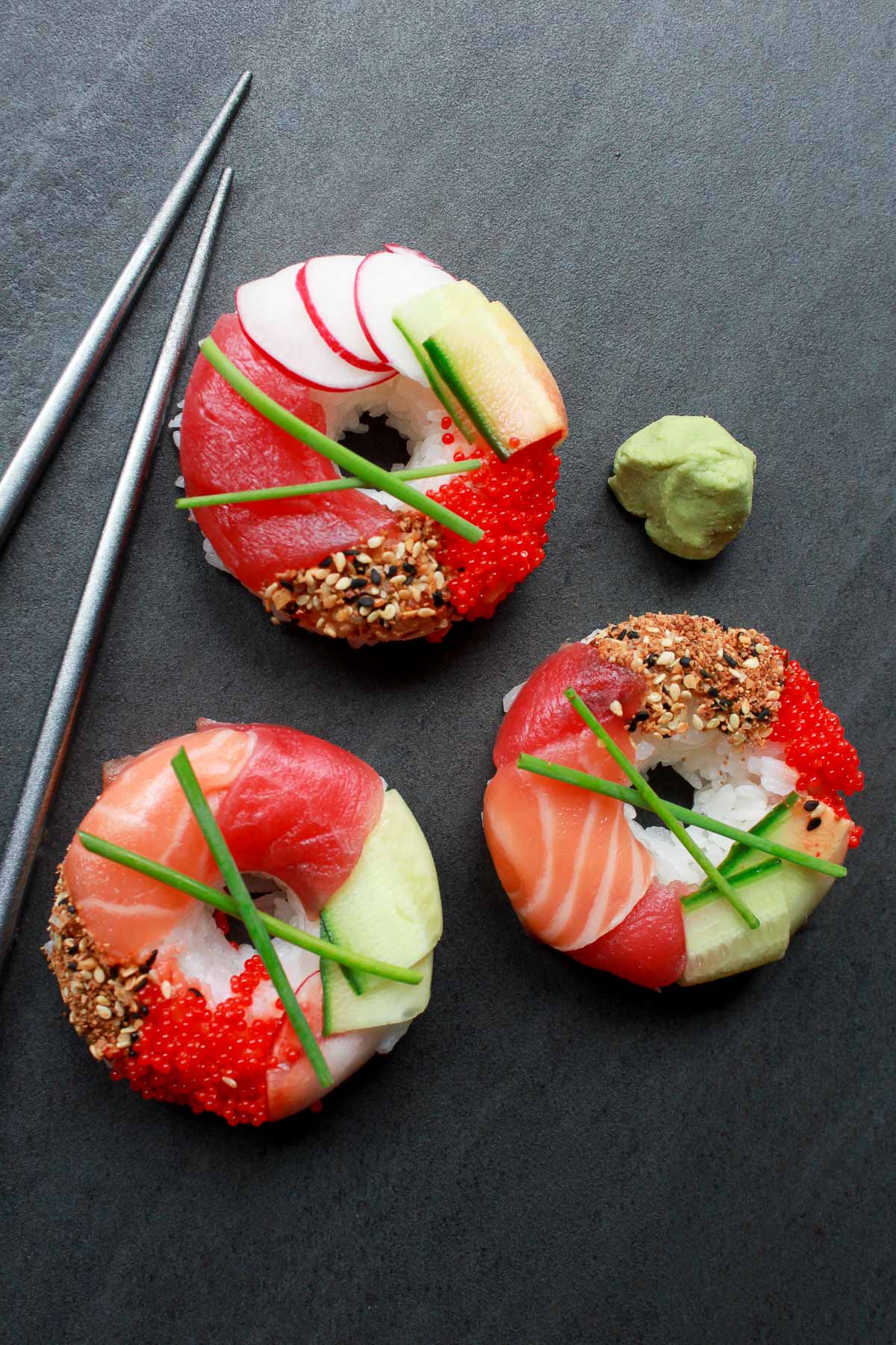Three sushi donuts on a black surface with silver chopsticks and wasabi paste on the side.