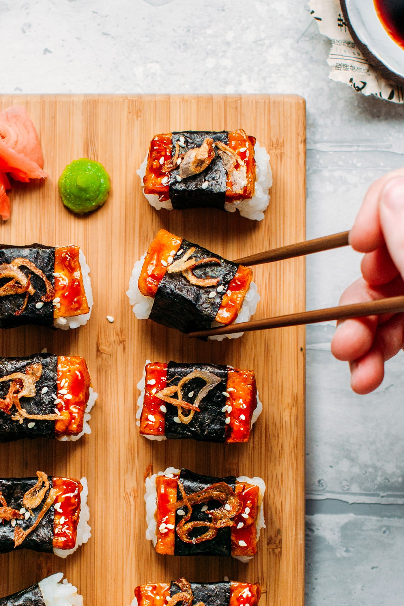 Teriyaki tofu nigiri pieces on top of a wooden board with a hand picking up a piece with wooden chopsticks.