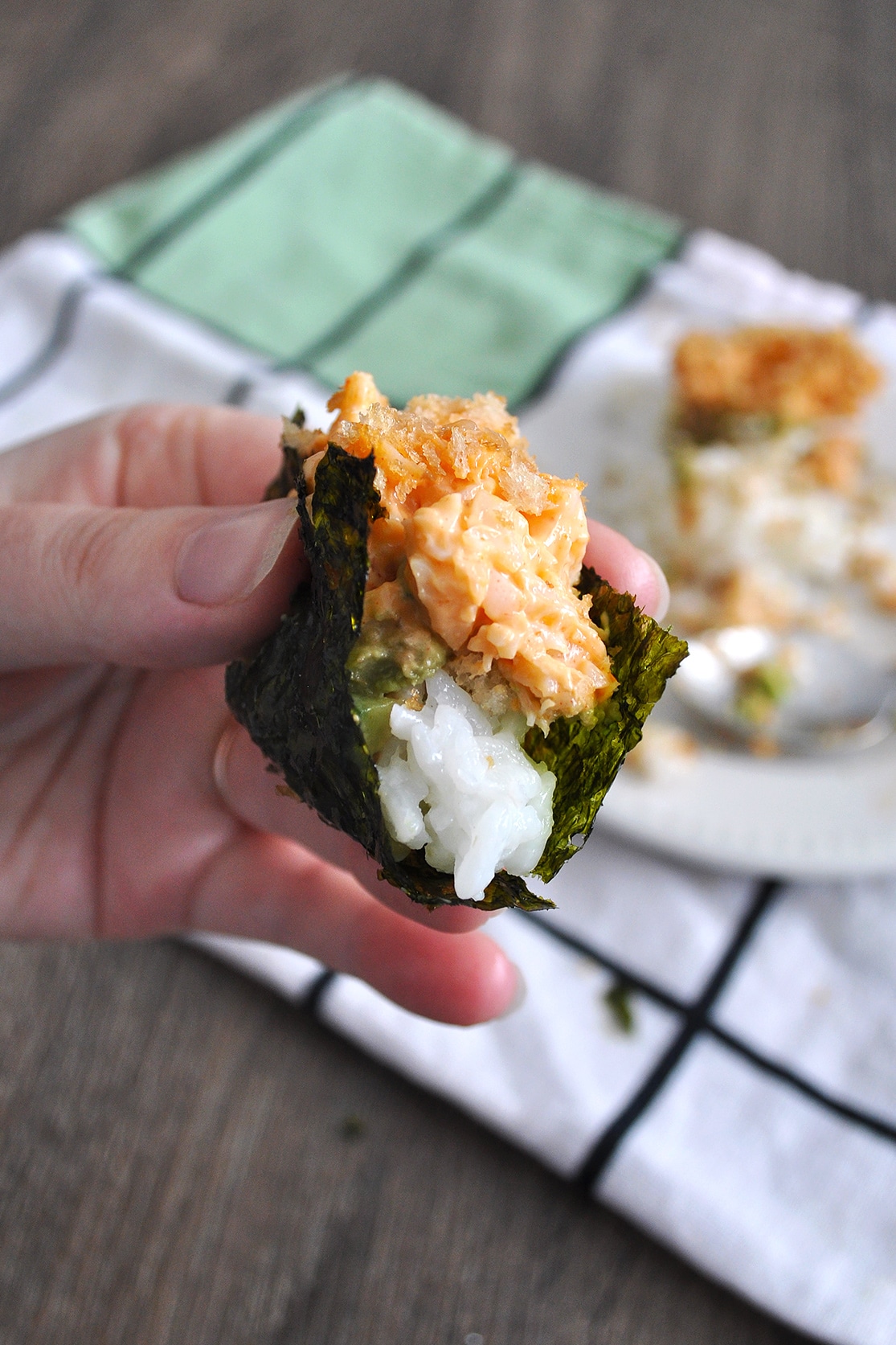 A hand holding up a piece of crispy shrimp pan sushi over a plate of sushi on top of a napkin.