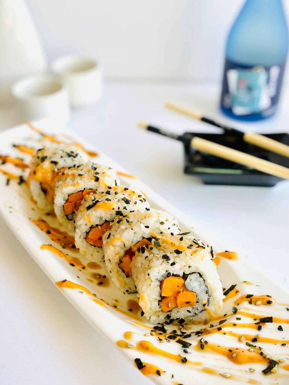 A sliced sweet potato veggie sushi roll on a narrow white plate with chopsticks and sake on the side.