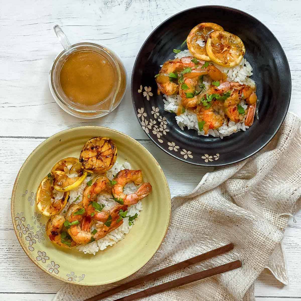 Two plates of grilled shrimp on top of white rice with charred lemon slices and miso sauce and chopsticks on the side on top of a white wooden board.