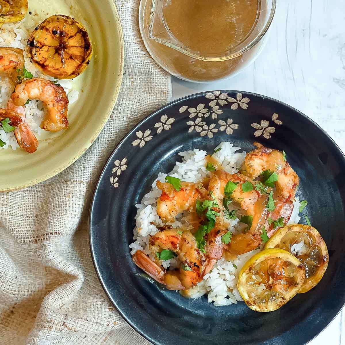 Two plates of grilled shrimp on top of white rice with charred lemon slices and miso sauce on the side on top of a white wooden board.