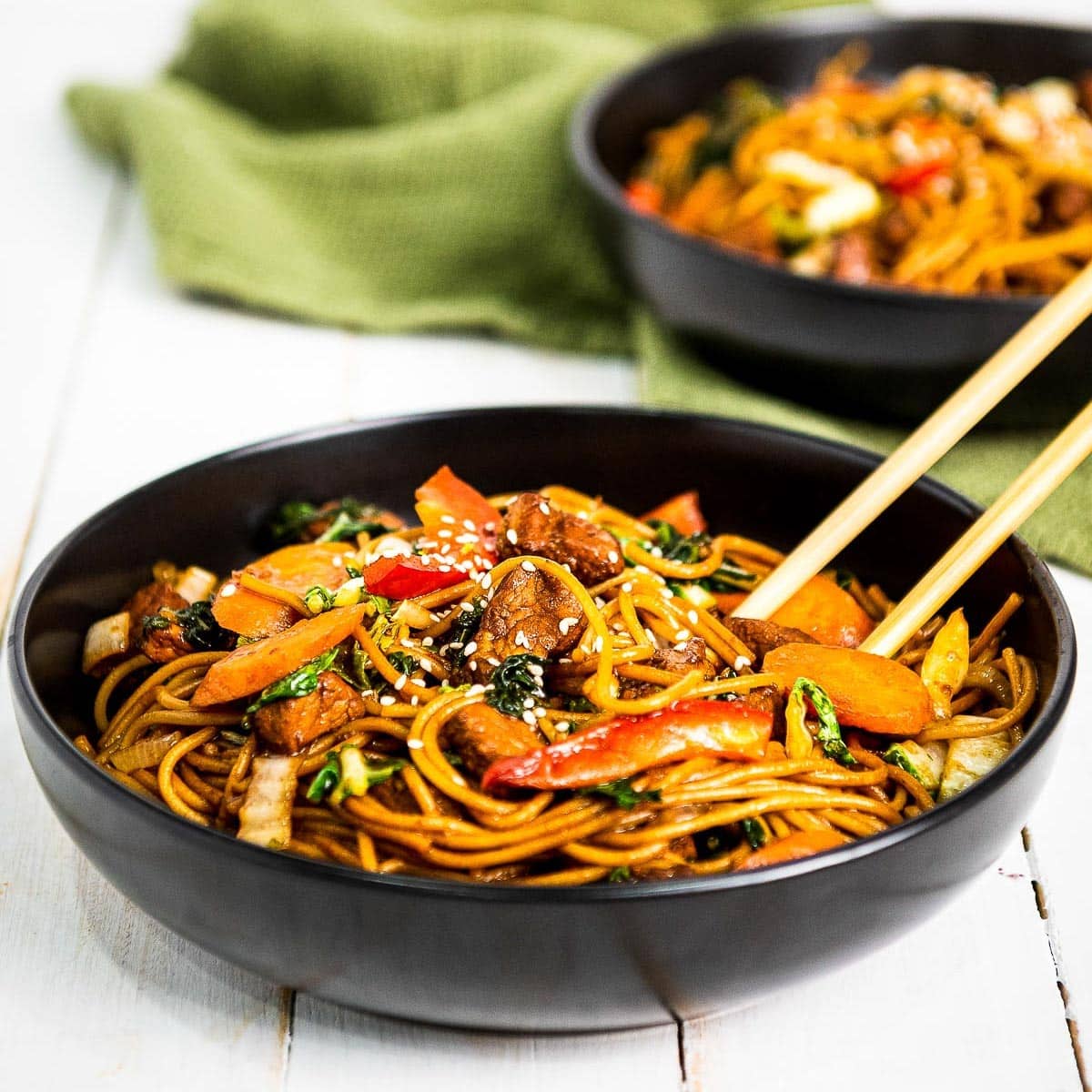 Pork Lo Mein in two black bowls with chopsticks and a green napkin on top of a white board.