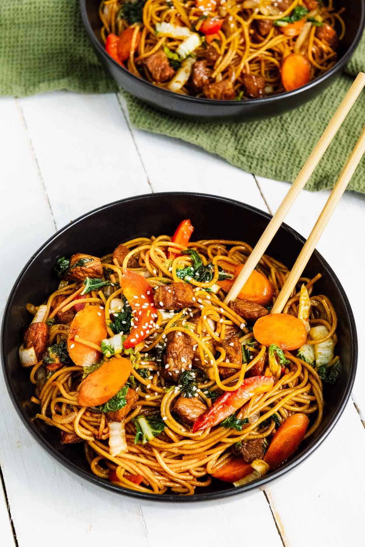 Pork Lo Mein in two black bowls with chopsticks and a green napkin on top of a white board.