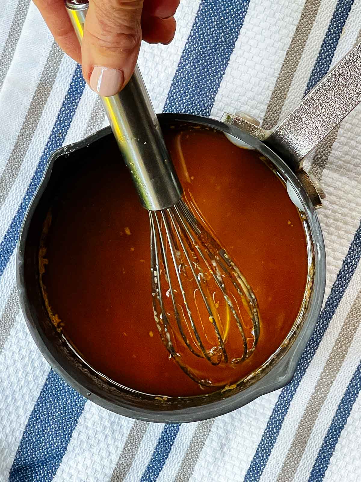 A hand with a whisk stirring a sriracha sauce mxture in a small sauce pan on top of a striped kitchen towel.