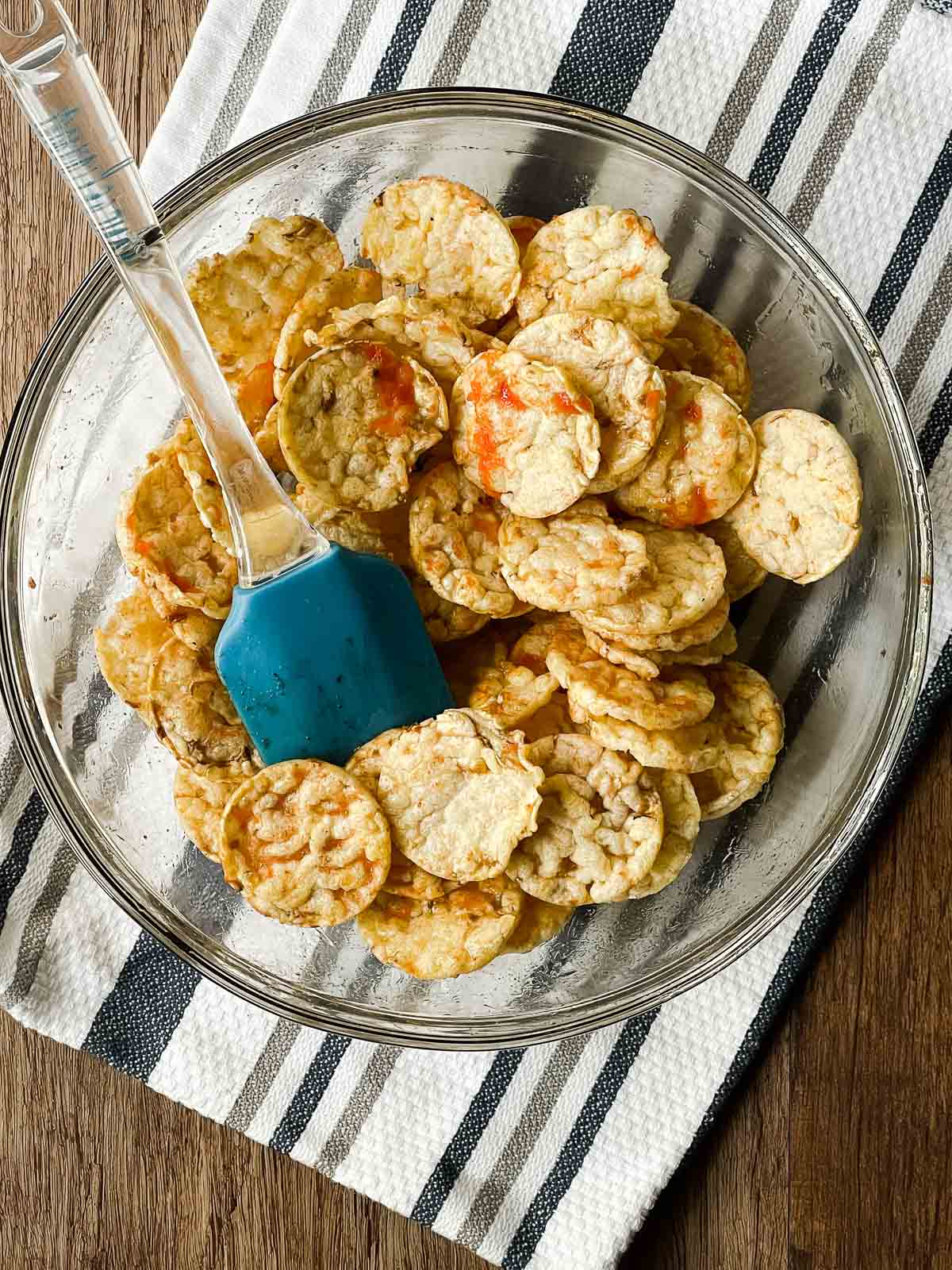 A blue silicone spatula stirring sriracha popcorn cakes in a glass bowl on top of a striped kitchen towel.