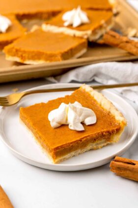 A piece of pumpkin pie topped with whipped cream on a white plate with a gold fork and a baking tray with other pieces in the background.