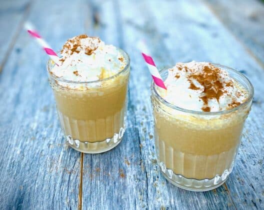 Two pumpkin pie milkshakes in glasses topped with whipped cream with a striped straw inserted, placed on top of a blue wooden board.