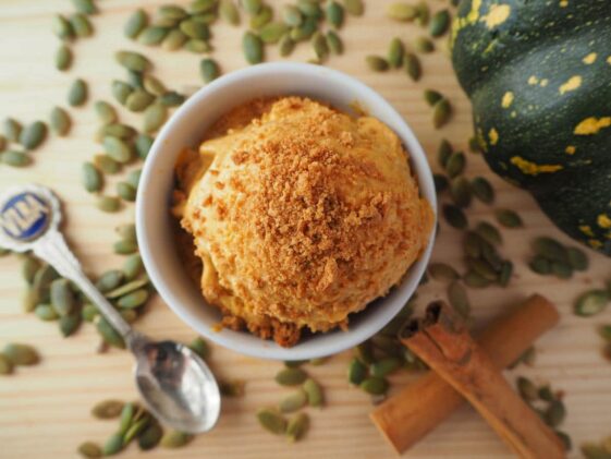 A scoop of pumpkin spice ice cream topped with cookie crumble in a white bowl with a spoon on the side.