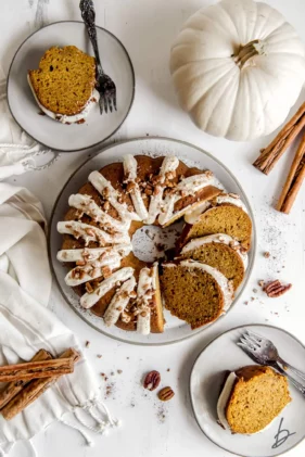 A beautiful pumpkin bundt cake with cream cheese frosting on a round plate with a slice on the side and a large white pumpkin in the background.