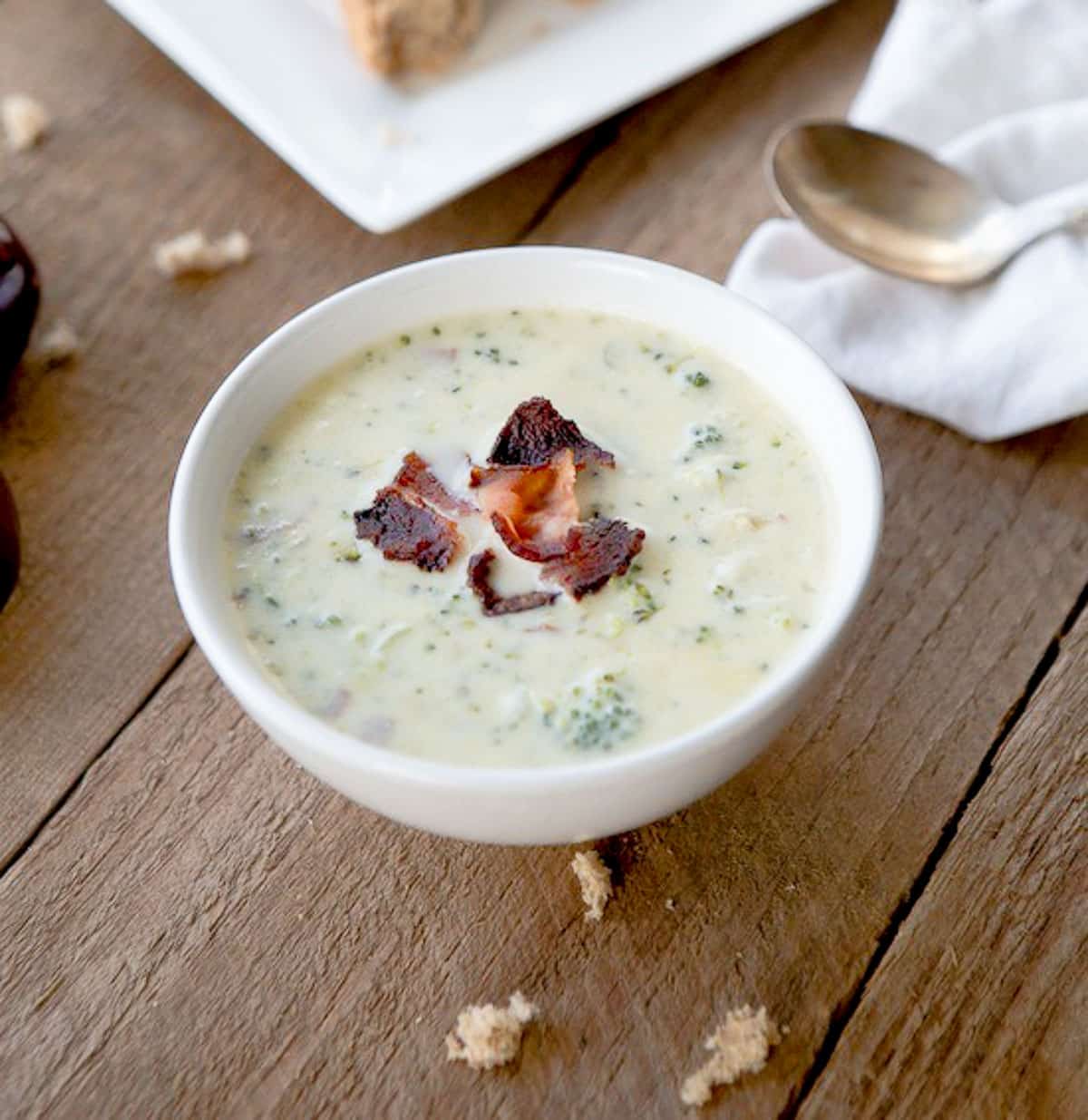 A white bowl of rich and hearty cream of broccoli soup with bacon bits on top placed on top of a wooden board.