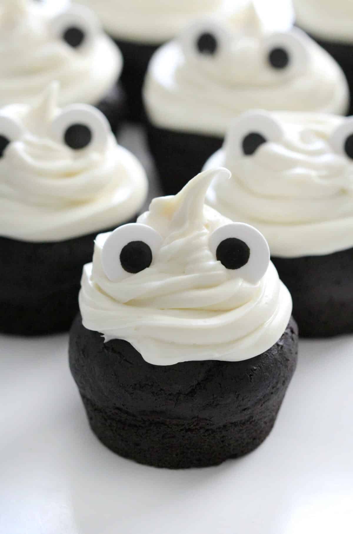 chocolate ghost cupcakes for Halloween decorated with vanilla frosting and candy eyeballs on a white surface