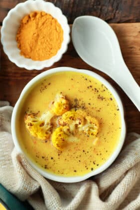 vibrant cauliflower soup in a white bowl with a white spoon and turmeric powder on the side