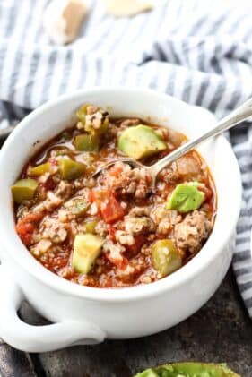 Stuffed pepper soup with chunks of avocado in a white soup bowl with a silver spoon inside and a striped napkin on the side.