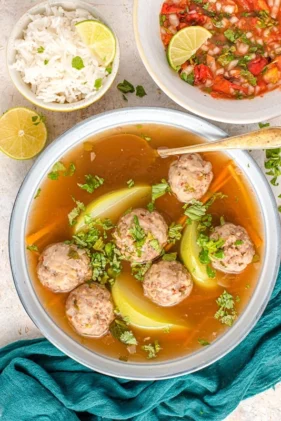 Meatball soup in beef broth served in white soup bowl with salsa and rice on the side.