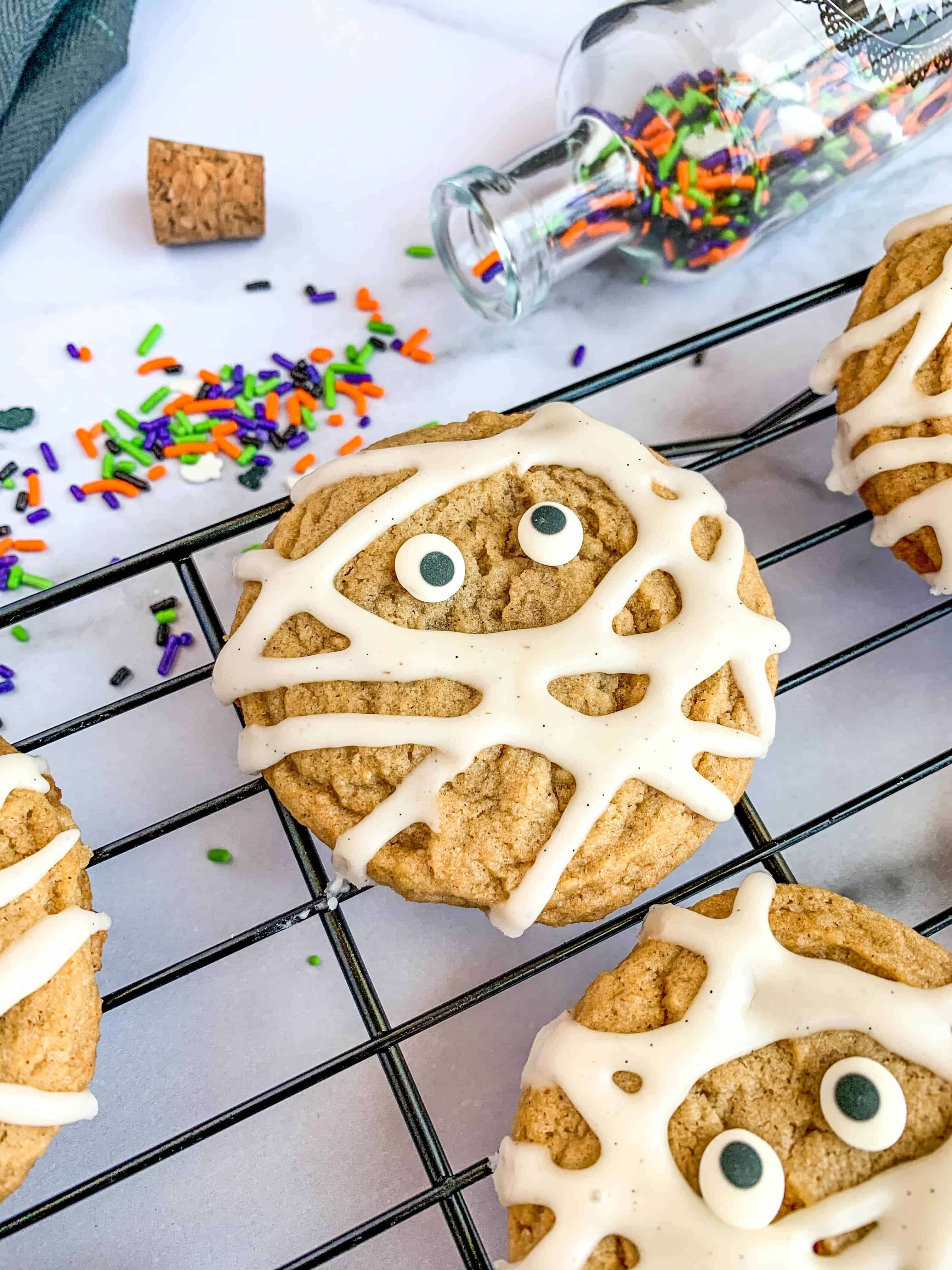 maple cinnamon cookies for Halloween decorated like mummies with eyeballs on top of a baking rack and colorful sprinkles on the side