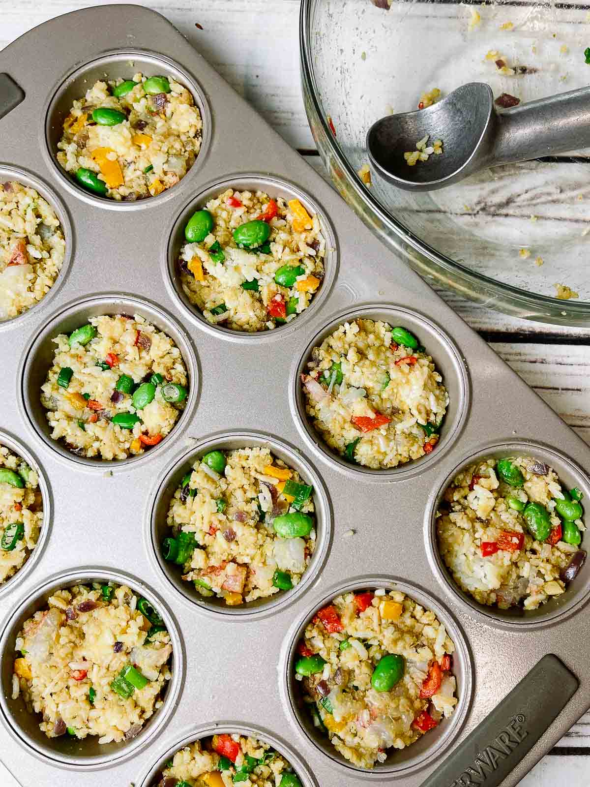 fried rice cups in a muffin tin ready for the oven with an empty glass mixing bowl in scooper on the side