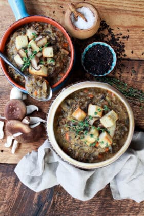 two bowls of wild rice mushroom soup topped with bread croutons on top of a wooden board with wild mushroom and uncooked wild rice on the side.