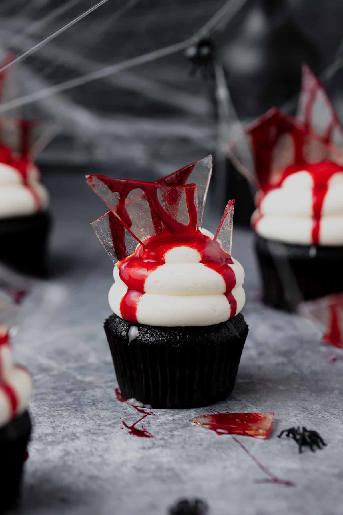 Bloody broken glass chocolate cupcakes for Halloween with vanilla frosting and topped with candy glass dripping with blood on top of a gray surface.