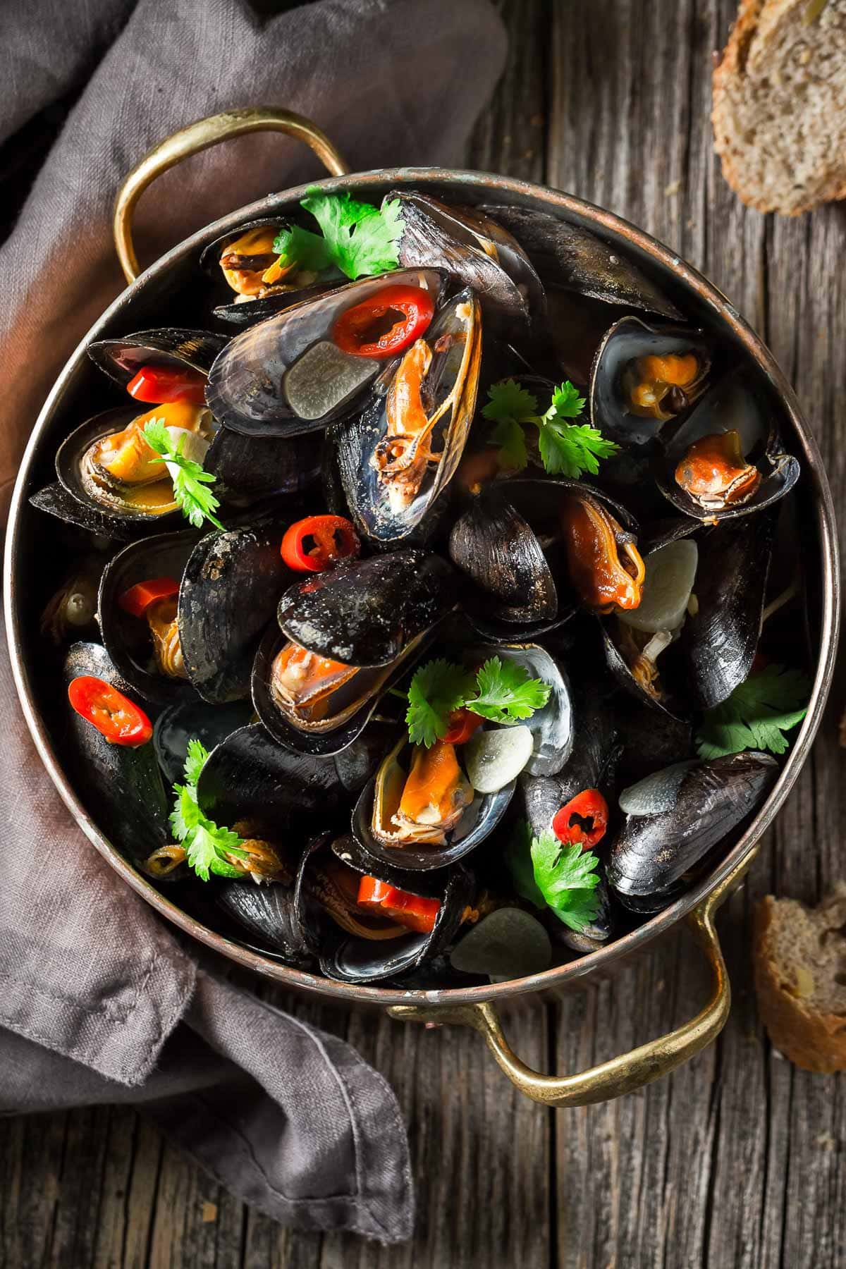 An overhead shot of a copper pot filled with steamed mussels garnished with red chili peppers and cilantro on a wooden board with sliced bread and a gray napkin on the side.