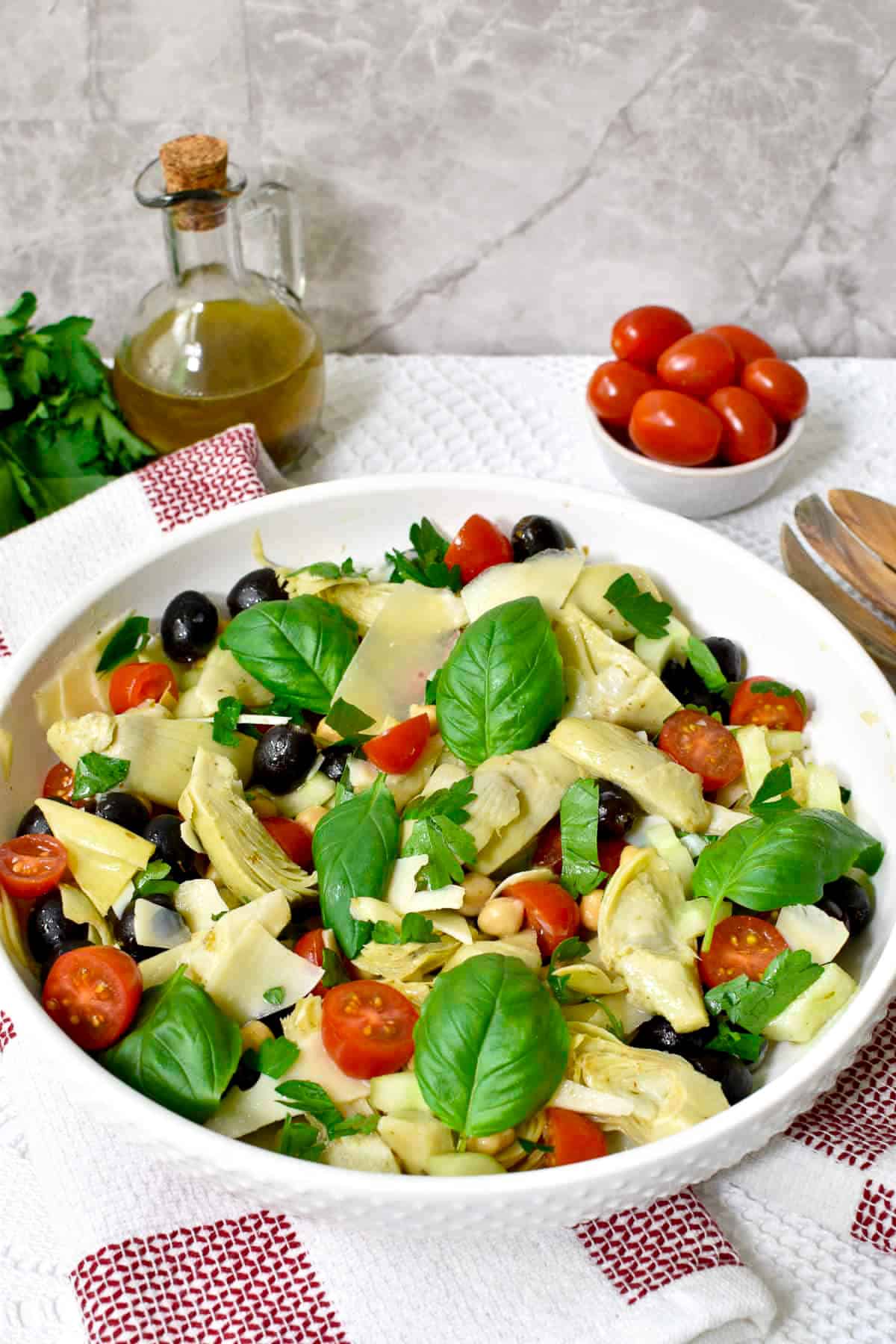 A round white bowl filled with artichoke salad with tomatoes, black olives, chickpeas, and topped with fresh basil leaves, and a small white bowl of cherry tomatoes on the side.