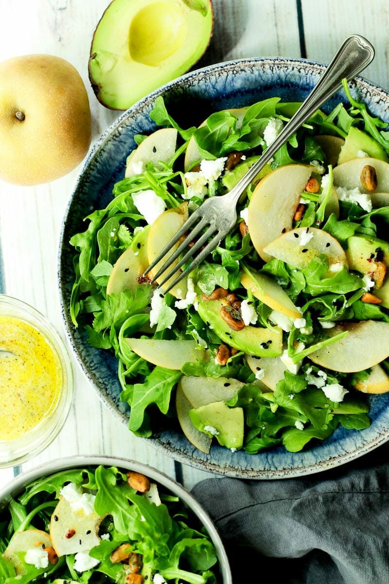 An arugula salad with sliced Asian pears and avocado in a blue bowl with a fork on top and half an avocado, whole pear, and lemon vinaigrette dressing on the side.