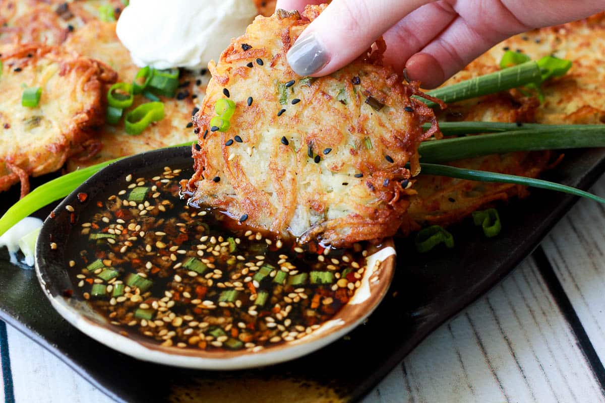 A hand dipping a Hanukkah side dish potato latke into an Asian soy dipping sauce in a small bowl, with more potatoes latkes on the side.
