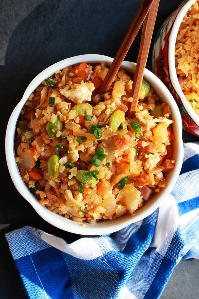 Cauliflower kimchi rice in a white bowls with brown chopsticks inserted and a blue and white checkered napkin on the side.