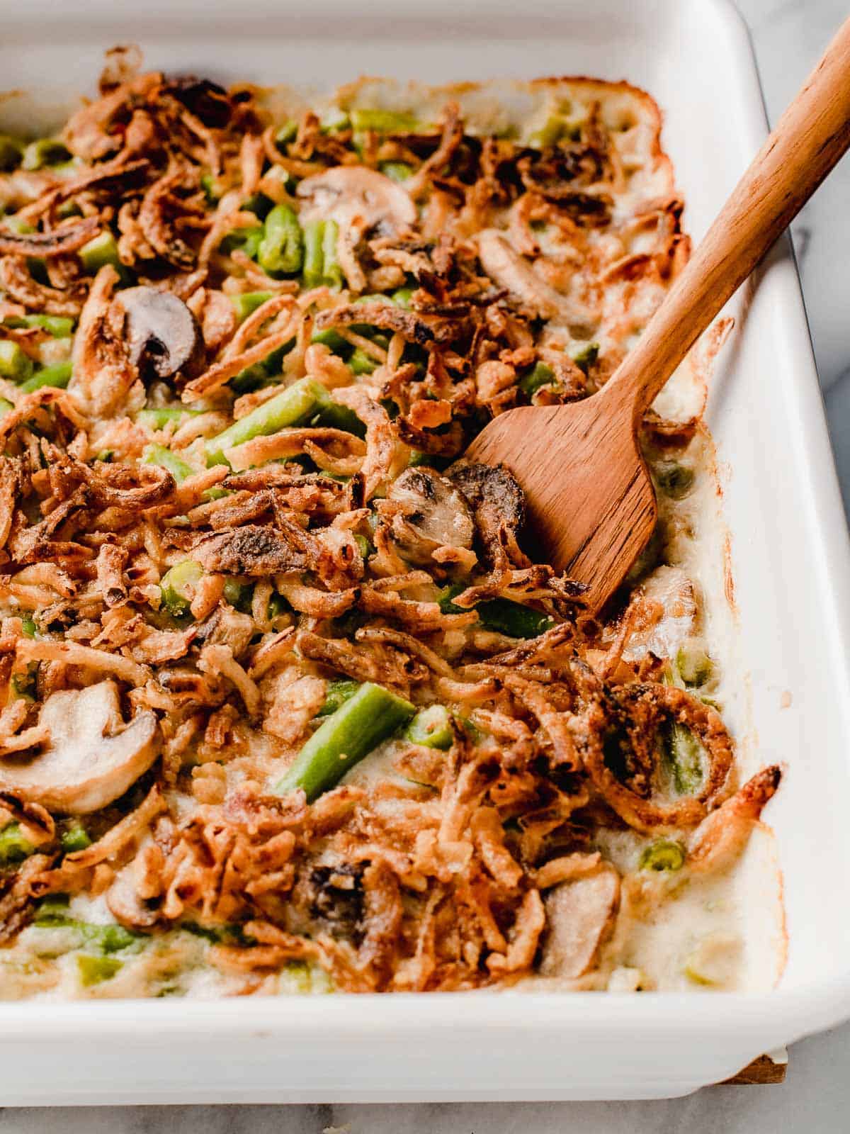 Green bean casserole side dish in a white serving dish topped with fried onions and a wooden spoon in the dish.