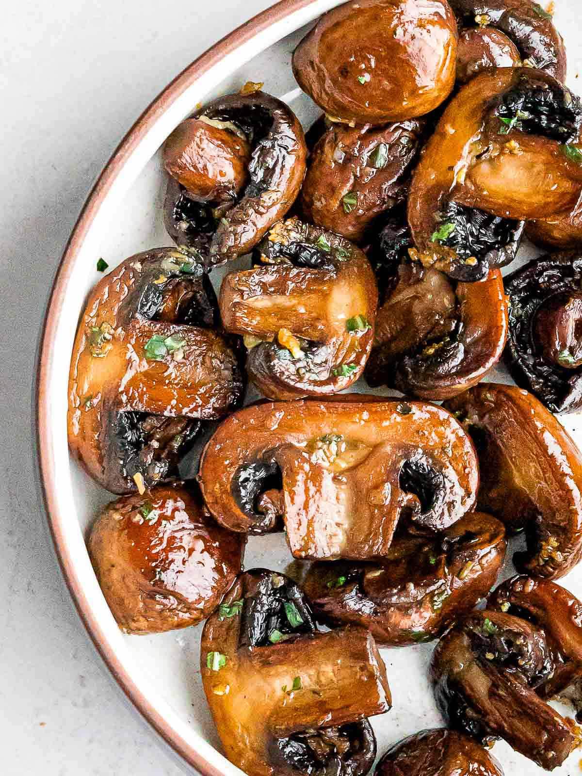 Sliced sautéed mushrooms side dish tossed in garlic butter topped with herbs on a round white plate.