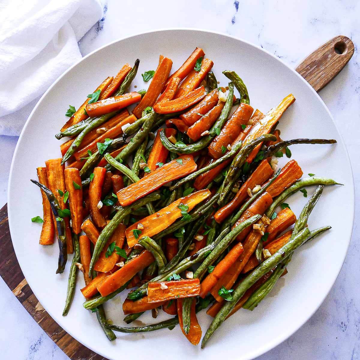 Roasted sliced carrots and roasted green beans on a large round white plate on top of a wooden board.