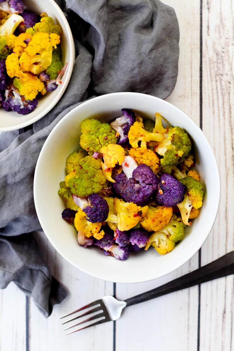 Cooked purple, yellow, and green cauliflower tossed in a sweet and spicy sauce, placed in a white bowl with a fork and gray napkin on the side on top of a white wooden board.