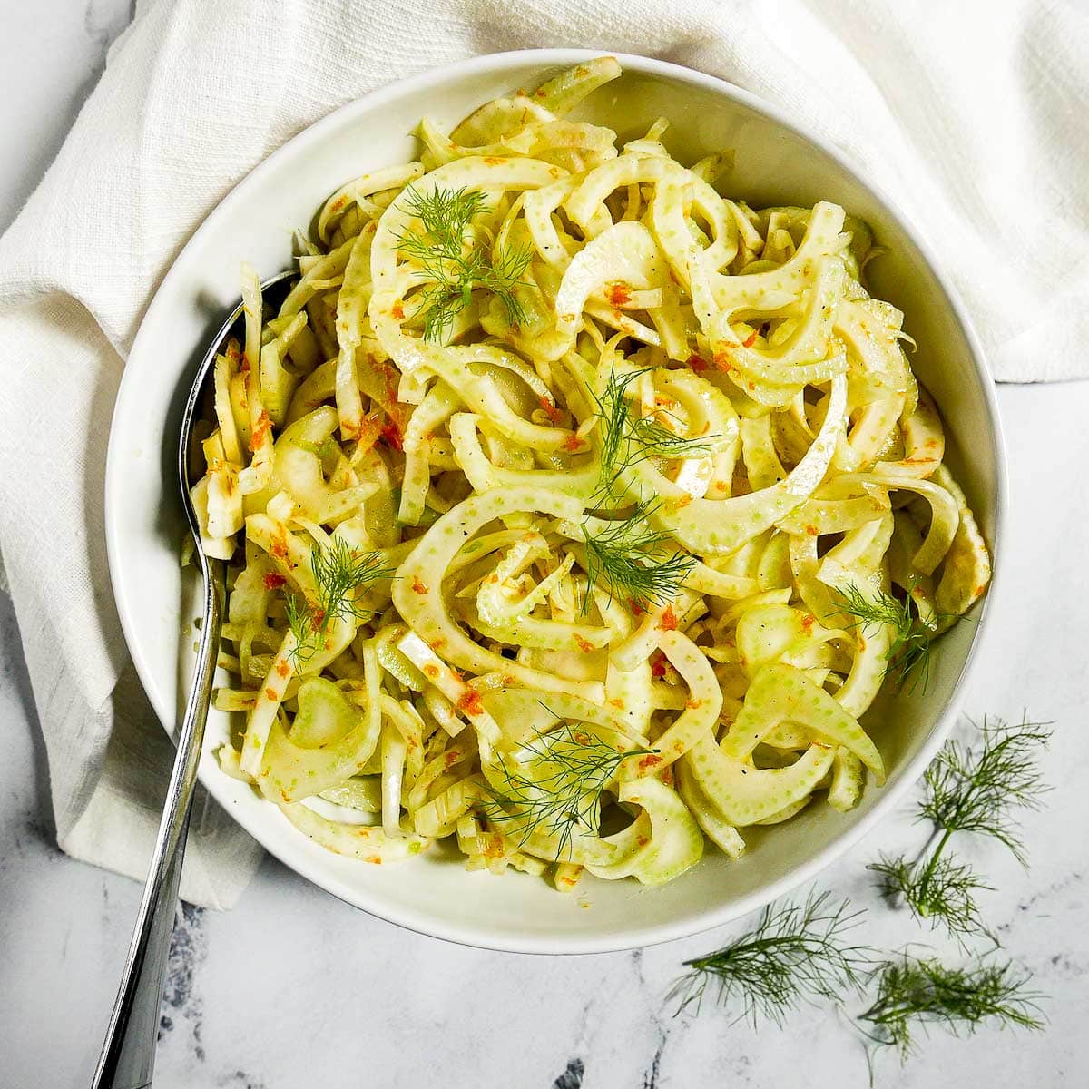 A shaved fennel salad topped with fennel fronds in a large white bowl with a large spoon inside on top of a white linen napkin.