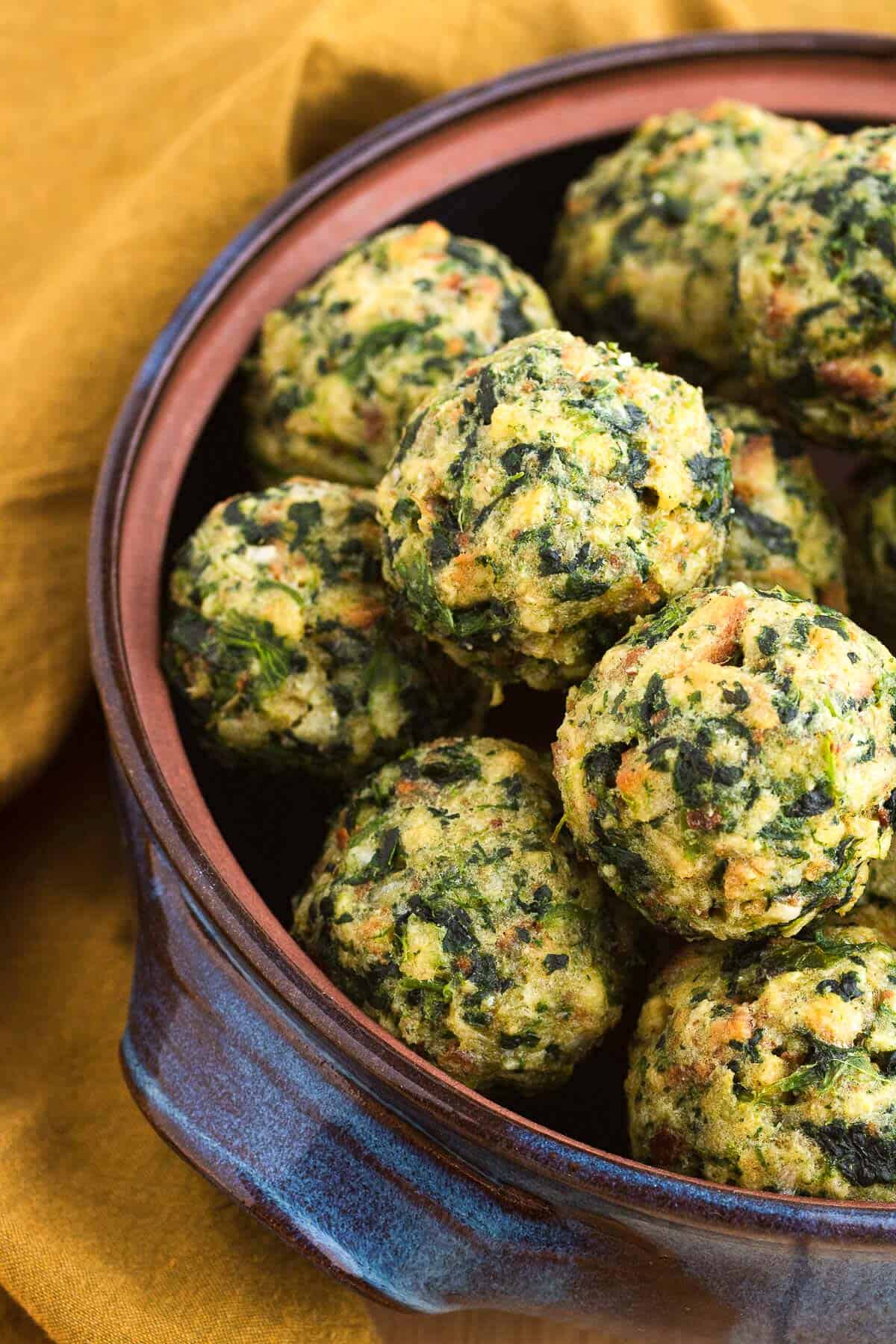 Baked spinach stuffing balls piled in a round blue bowl on top of a yellow linen napkin.