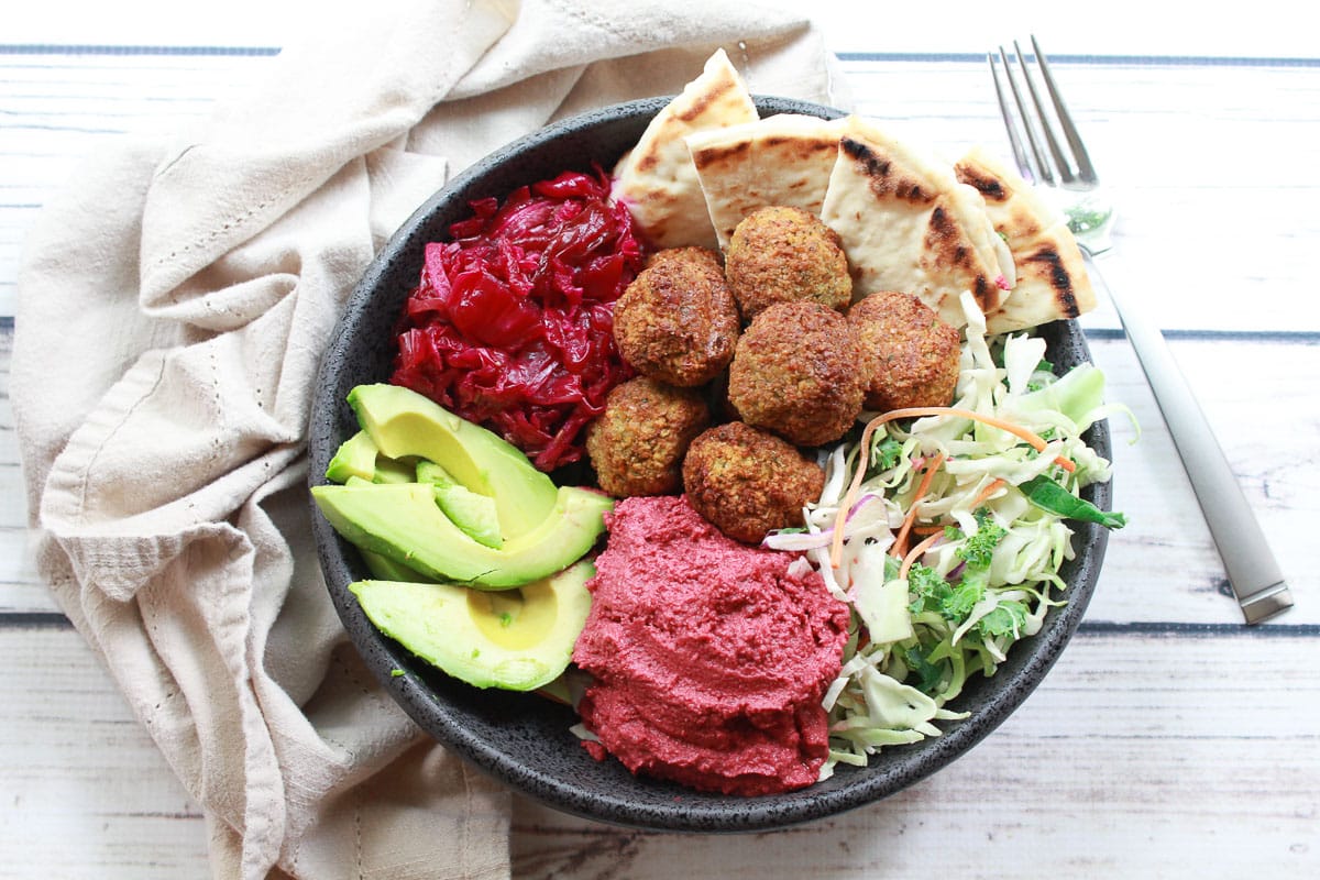 A large bowl with air-fried falafel balls, red beet hummus, pickled red onions, sliced avocados, mixed greens, and slices of pita bread with a fork and napkin on the side.