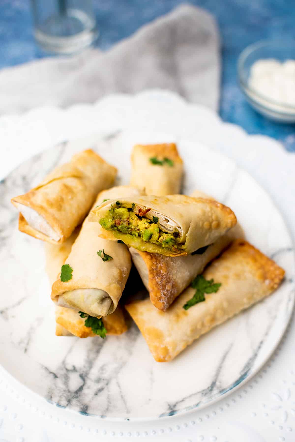 Vegan Avocado Egg Rolls stacked on a white marbled plate on top of a blue board with a small bowl of sour cream in the background.