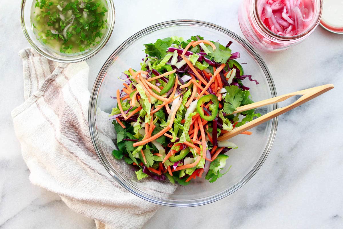 Fresh sliced vegetables in a glass bowl for making Nuoc Cham slaw with pickled red onions in a jar and nuoc cham on the side.