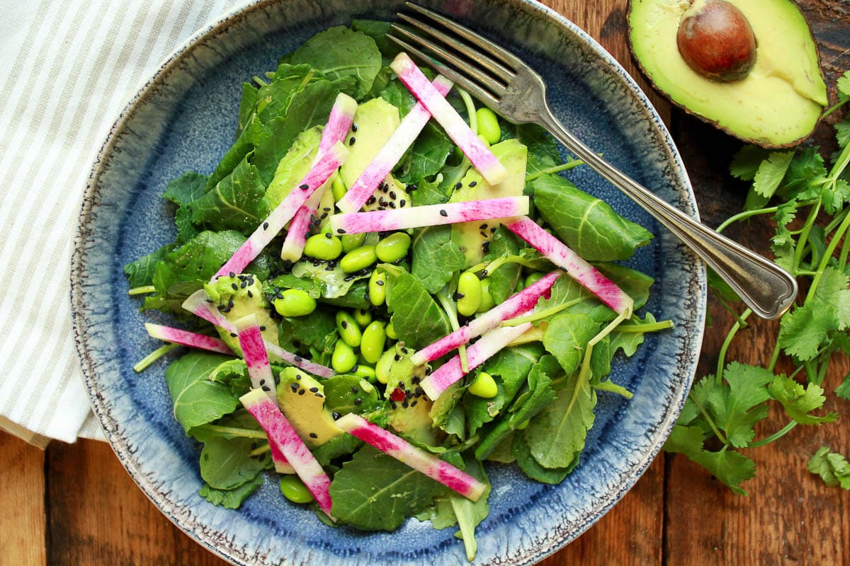 Avocado edamame kale salad in a blue bowl with a fork and sliced avocado and herbs on the side.