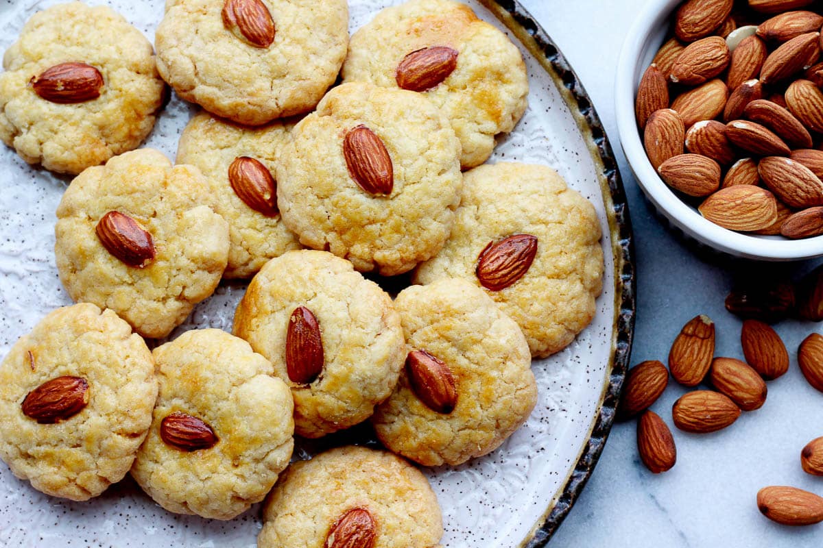 Chinese almond cookies stacked on a round plate with a bowl of raw almonds on the side.