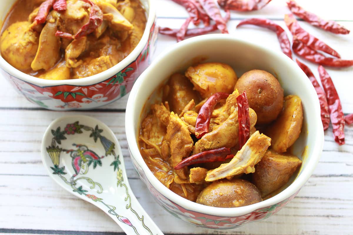 Two bowls filled with Chinese curry chicken and potatoes with whole red chili peppers on top all placed on a white wooden board with a white spoon on the side.