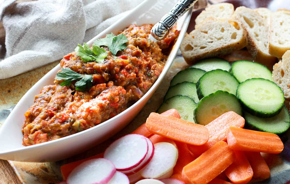 Roasted eggplant dip in a white narrow bowl with fresh veggies and bread slices on the side.