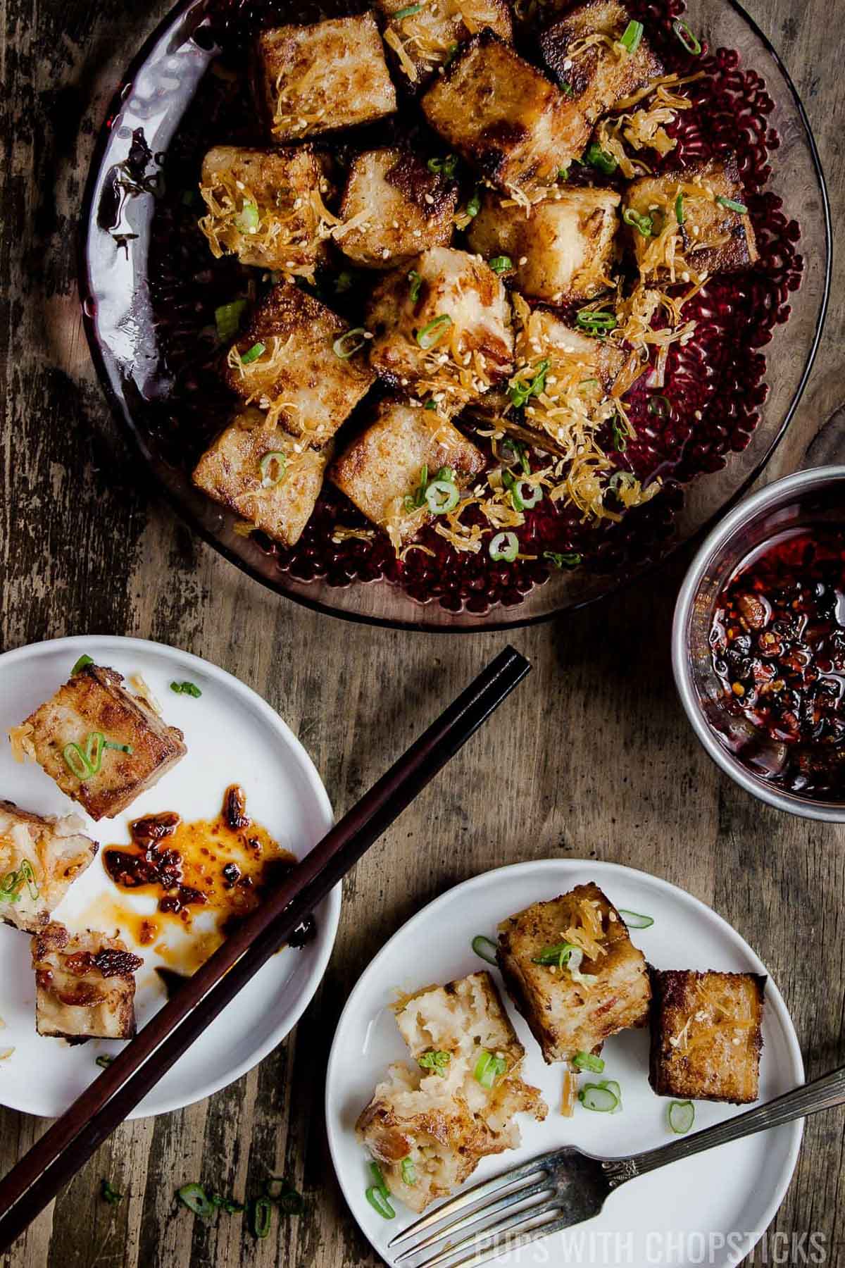 Cut up squares of Chinese turnip cake on various plates with black bean sauce in a bowl on the side on top of a wooden board.
